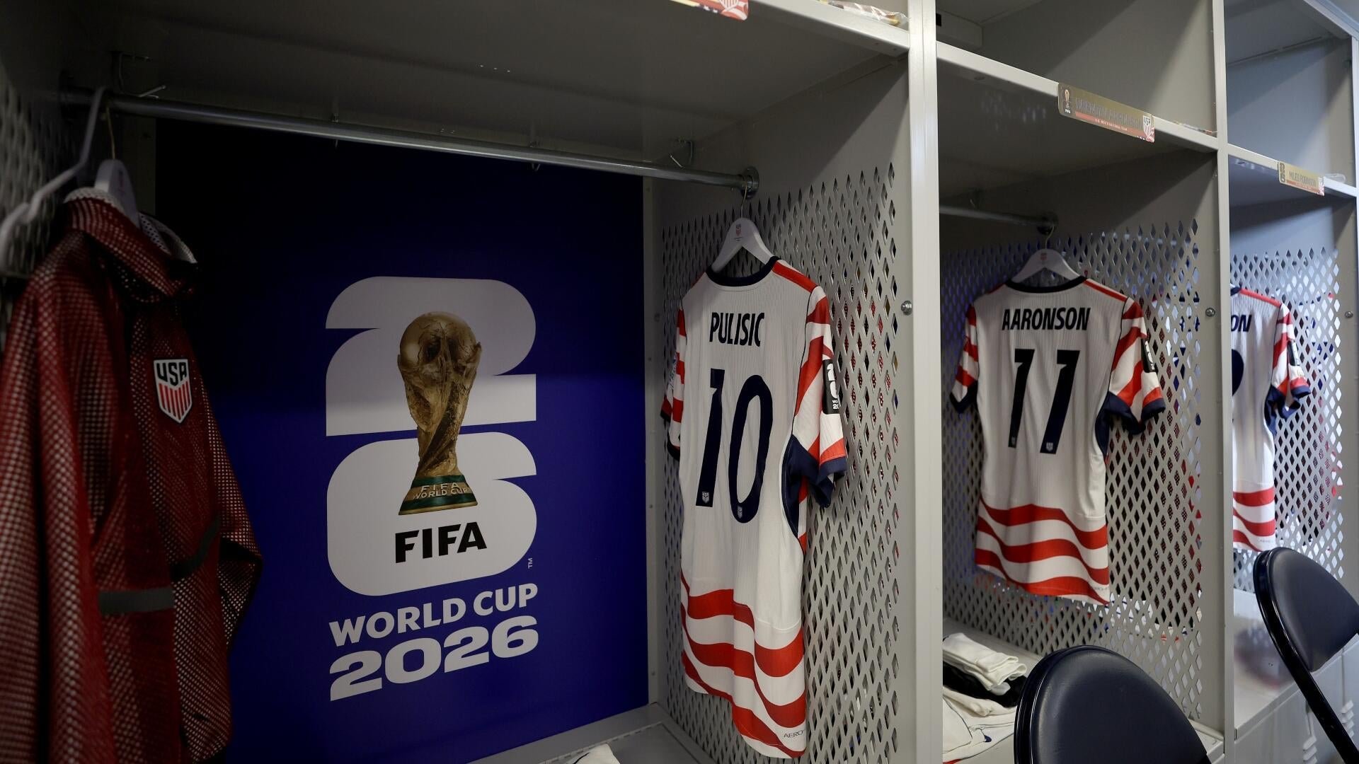 A locker room displays a FIFA World Cup 2026 sign alongside red, white, and blue USA jerseys labeled "Pulisic 10" and "Aaronson 11" hanging in open stalls.