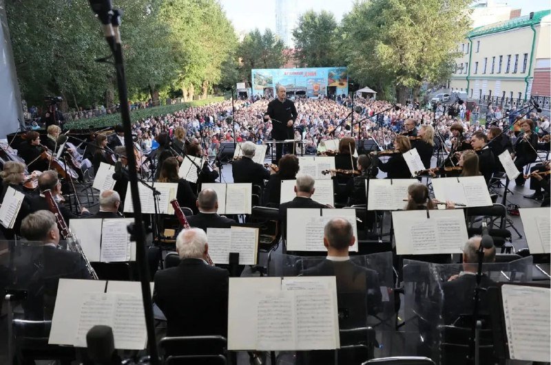 A symphony orchestra performs outdoors from a stage before a seated crowd of hundreds, with a digital billboard displaying "27 СЕНТЯБРЯ 2021" visible in the background.