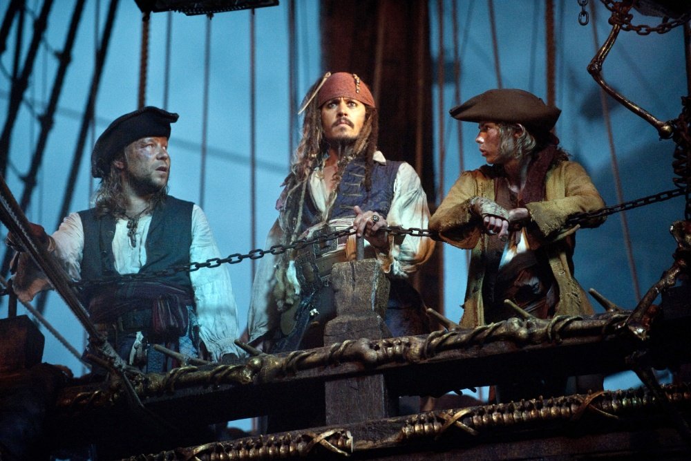 Three men in pirate costumes with tricorn hats stand on a wooden ship deck beside rigging and chains.