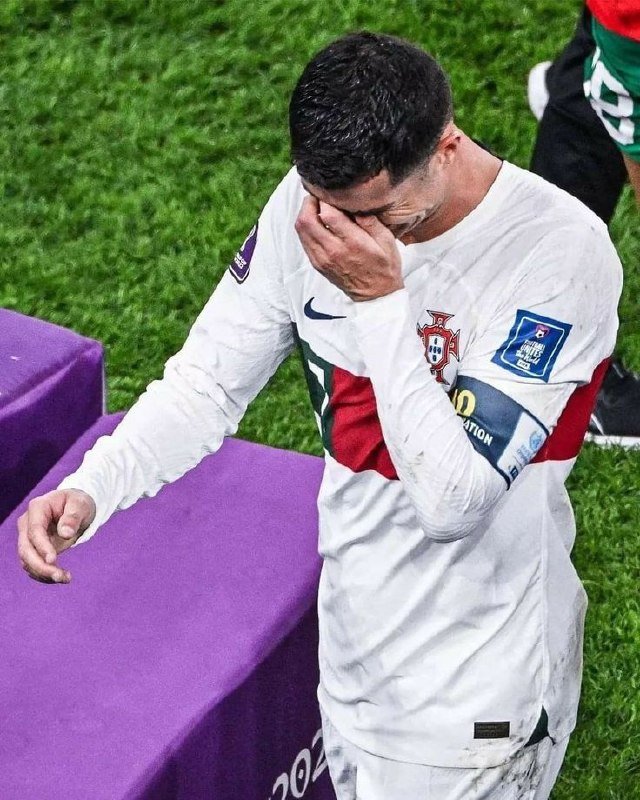 A soccer player wearing a white Portugal national team jersey with the captain's armband covers his face with his hand on the field after a match.