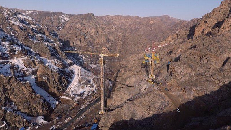 Two tall construction cranes stand among rugged, snow-dusted mountain slopes under a clear sky.