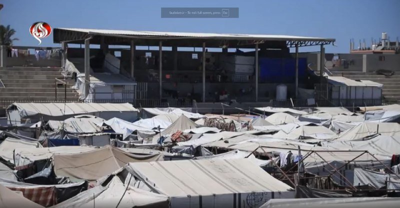 A densely packed encampment of white tents and tarps fills the foreground, set against a large covered stadium structure under a clear blue sky.