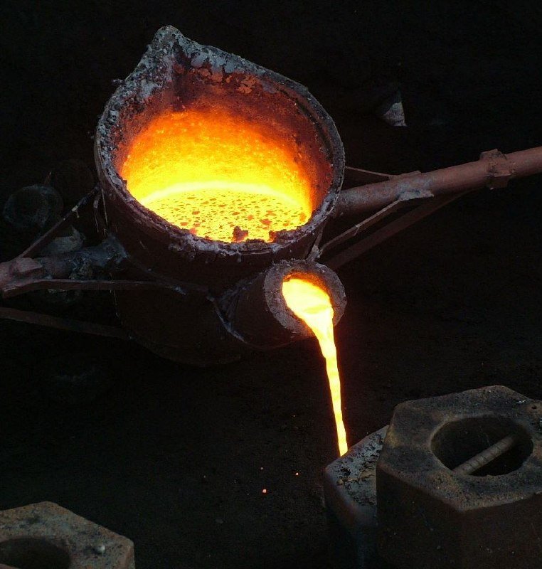 Molten metal glows bright orange as it pours from a worn industrial crucible into a hexagonal mold against a dark background.