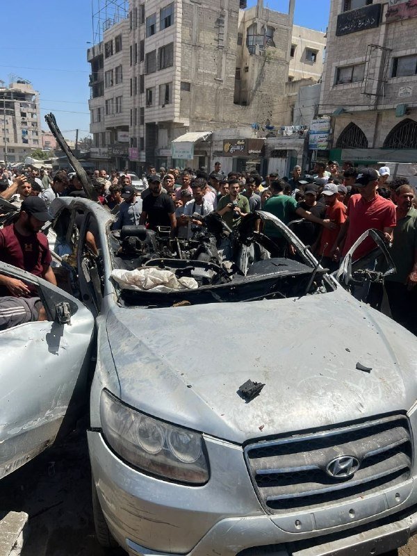 A crowd gathers around a heavily damaged silver Hyundai SUV with its windshield blown out and interior burned, set on a city street lined with multi-story buildings.