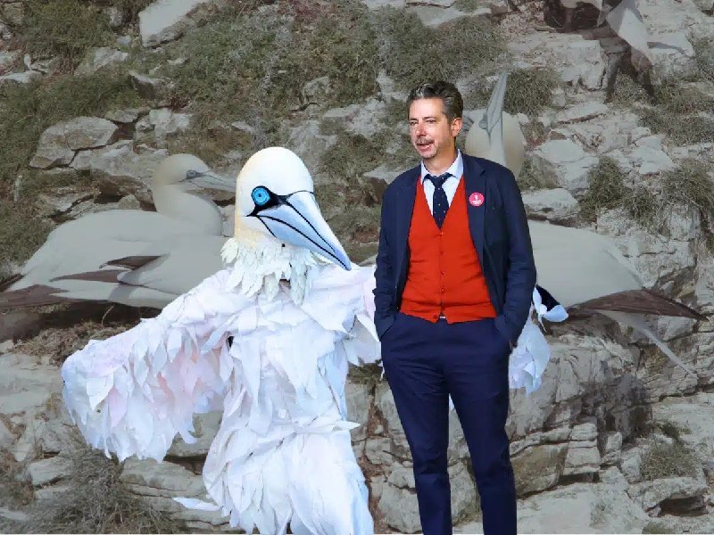 A man in a navy suit and red waistcoat stands beside a person in an elaborate white seabird costume against a rocky cliffside backdrop featuring real gannets.
