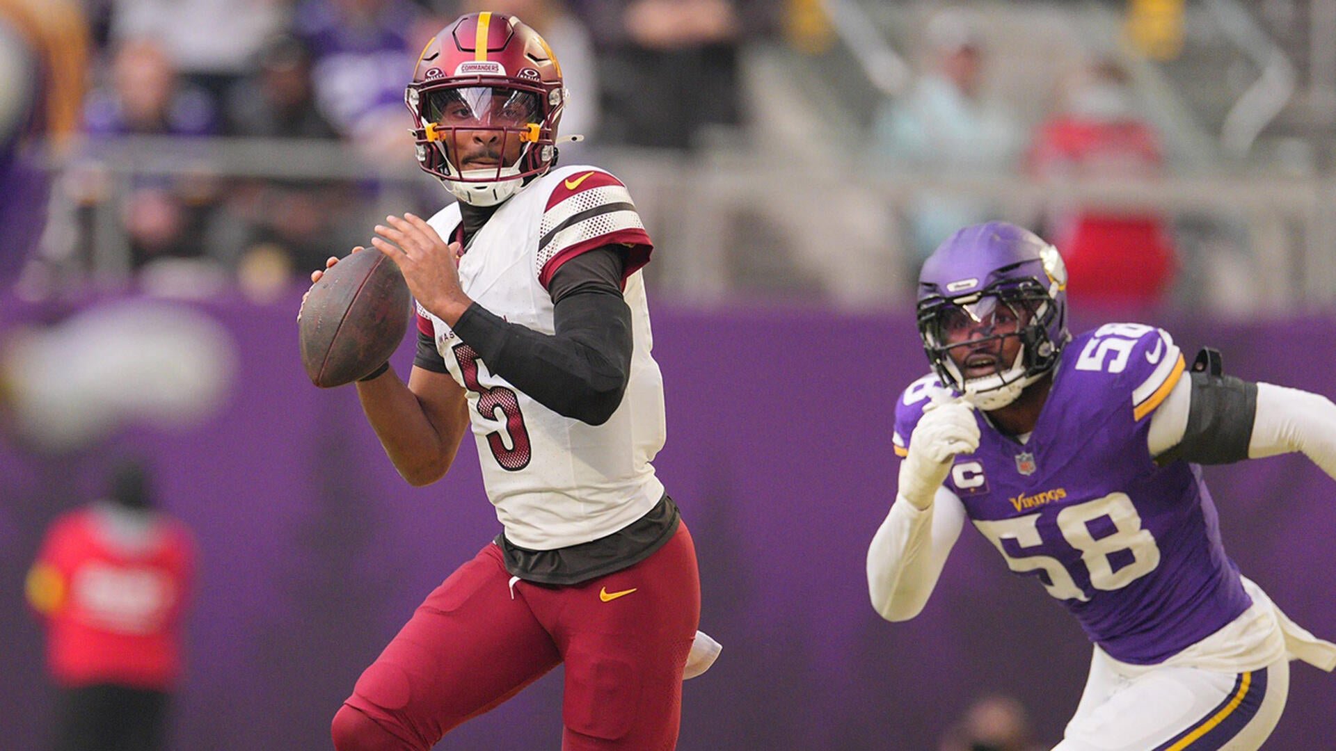 A quarterback in a white and burgundy uniform prepares to throw a football while a defender wearing a purple "Vikings" jersey numbered 58 rushes toward him.
