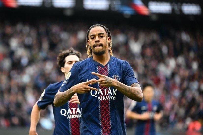A soccer player in a dark blue Paris Saint-Germain jersey with "QATAR AIRWAYS" branding points to his wrist in celebration, with a teammate and crowded stadium visible behind him.
