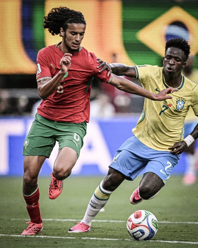 Two soccer players compete for a ball during a match, one wearing a red jersey with green shorts and the other in a yellow jersey with light blue shorts.