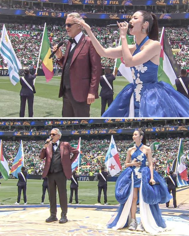 Two performers, a man in a maroon jacket and a woman in a blue gown, sing into microphones on a stadium field as flag-bearers display various national flags.