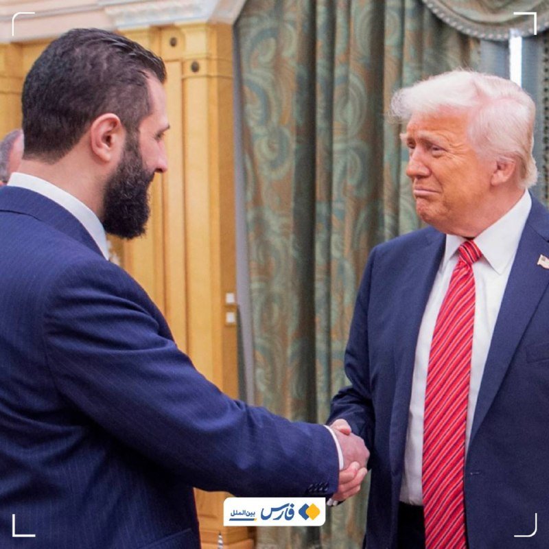 A bearded man in a dark suit shakes hands with an older man with white hair wearing a navy suit and red striped tie, indoors near a curtain.