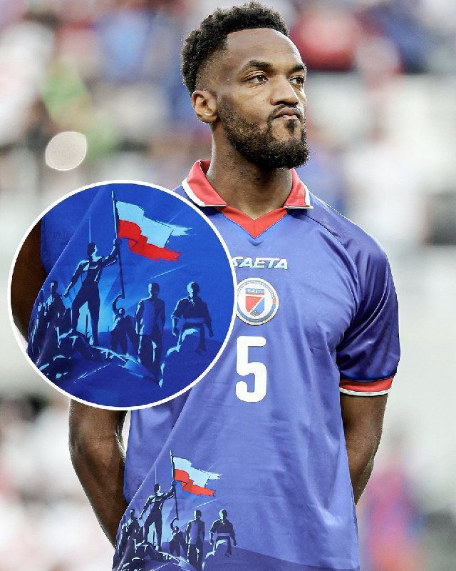 A bearded soccer player wearing a blue Haiti national team jersey with the number 5 stands on a field, the jersey featuring a circular graphic of figures holding a flag.