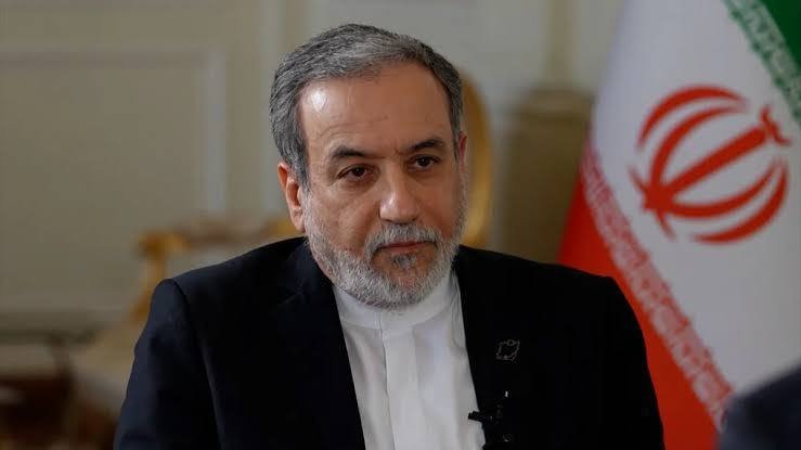 A gray-haired man in a dark suit and white shirt speaks while seated, with an Iranian flag visible behind him.