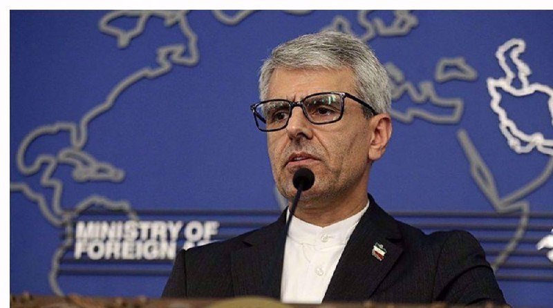 A gray-haired man wearing glasses and a dark suit speaks at a podium against a blue backdrop displaying a map outline and the text "MINISTRY OF FOREIGN."