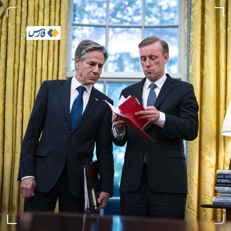 Two men in dark suits stand in a room with yellow curtains and a window, reviewing a red folder together, one holding a binder.