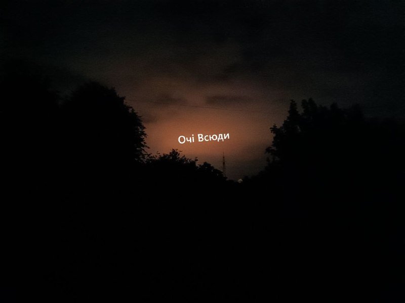 The illuminated Cyrillic text "Очі Всюди" glows against a dark, cloudy sky above silhouetted treetops at dusk.