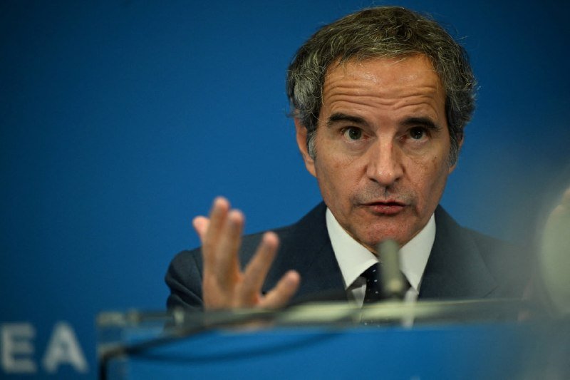 A man in a dark suit and patterned tie speaks behind a clear podium, gesturing with his hand against a blue backdrop displaying partial white lettering.