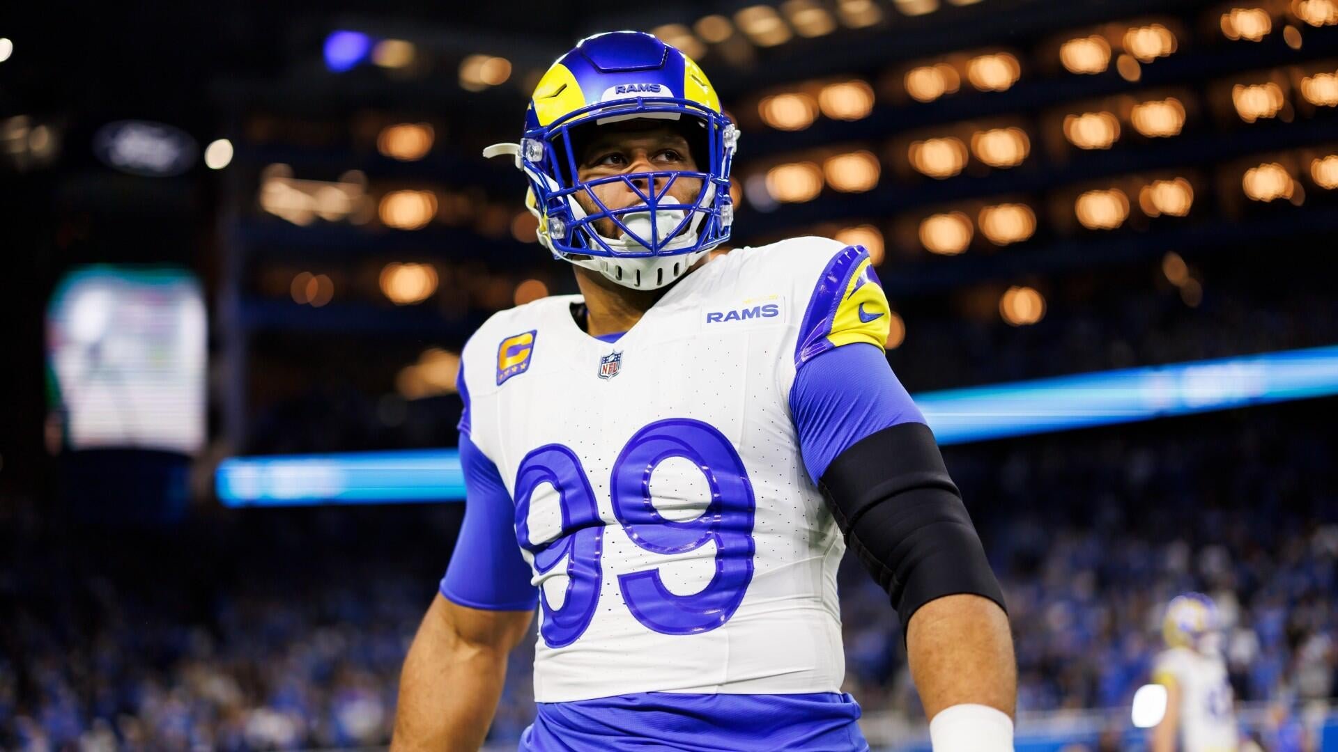 A football player wearing a white Rams jersey with the number 99 and a blue and yellow helmet stands on the field inside a domed stadium.