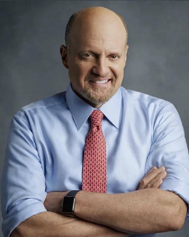 A bald man with a goatee, wearing a light blue dress shirt, pink patterned tie, and a smartwatch, stands smiling with arms crossed against a gray background.
