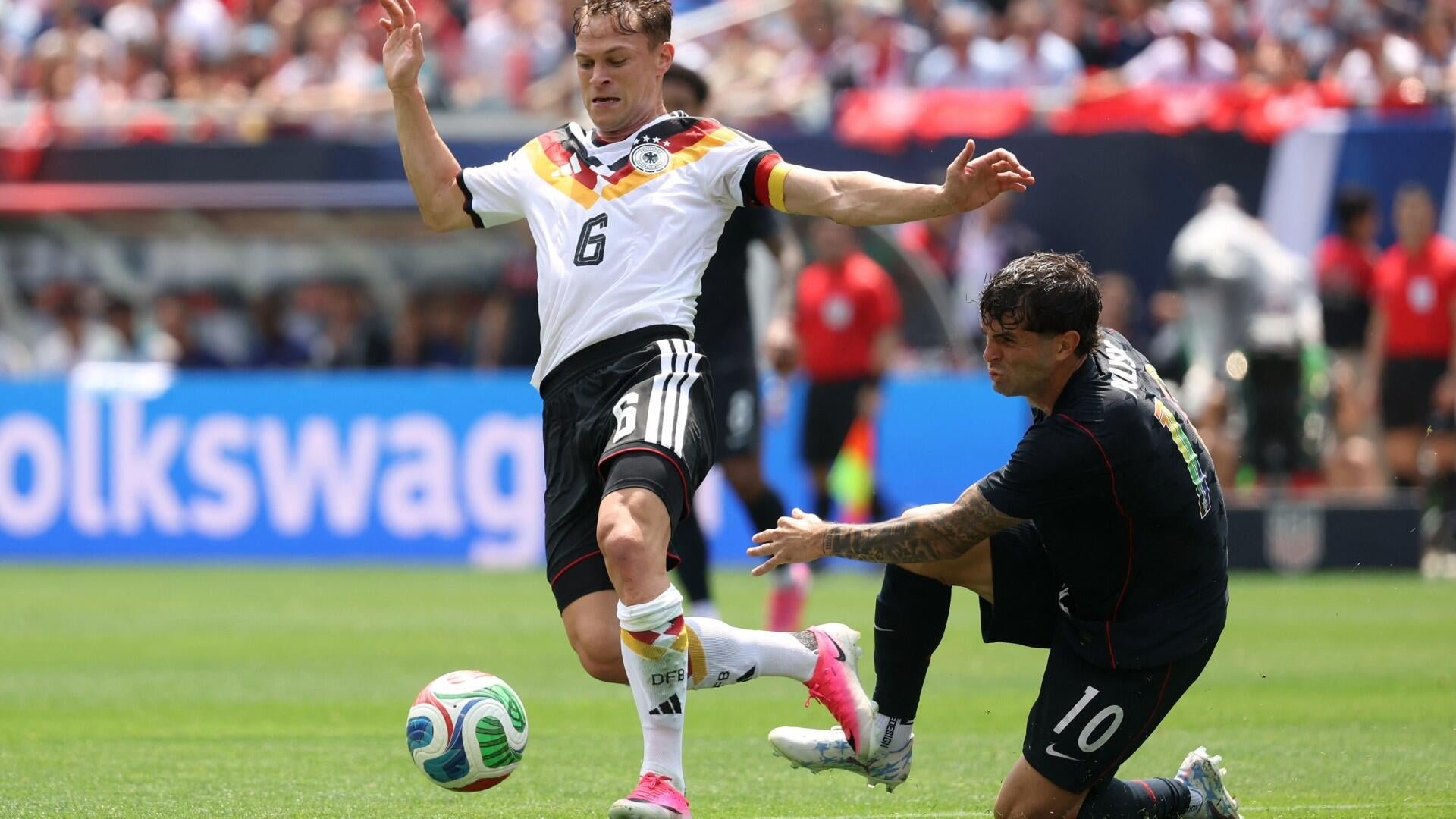 Two soccer players compete for the ball during a match, with one wearing a white Germany jersey (#6) and the other in a black Mexico jersey (#10), in a stadium filled with spectators.