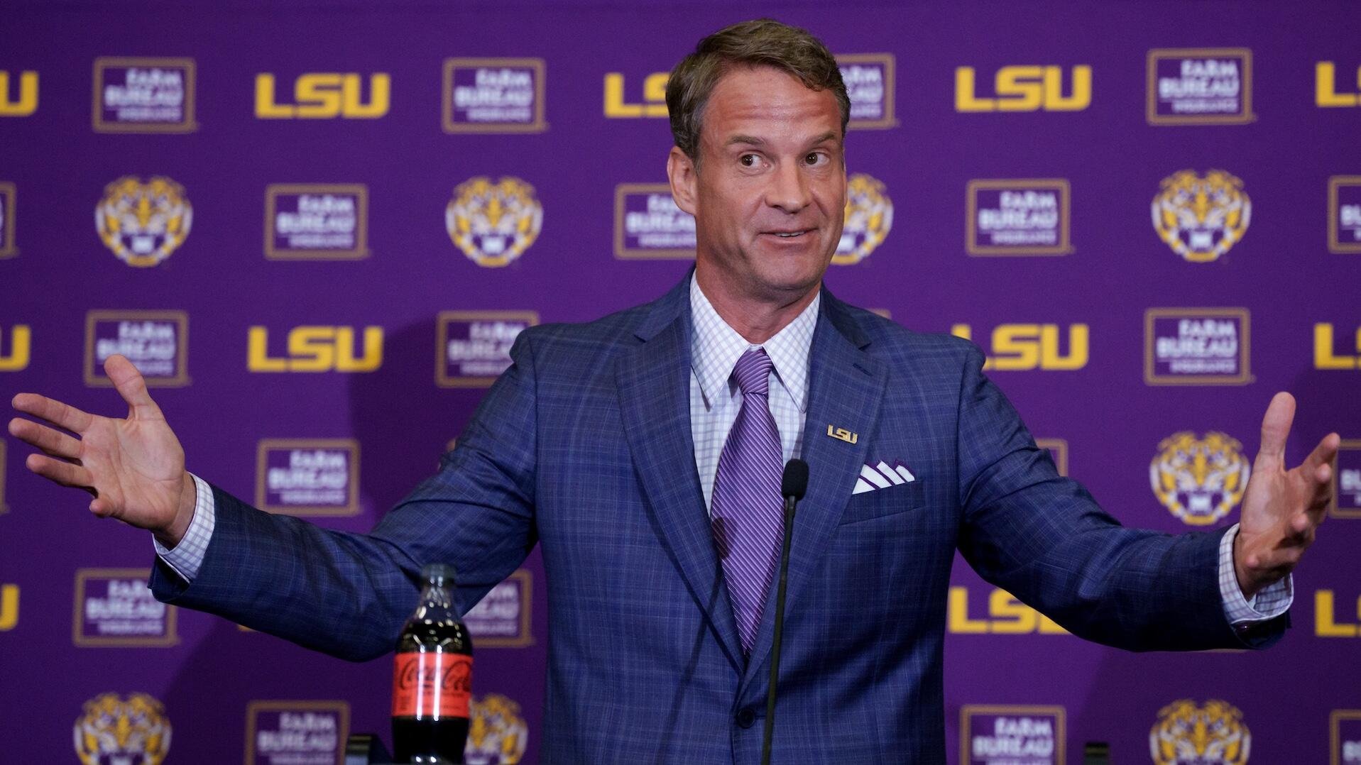 A man in a blue suit and purple tie stands at a microphone with arms outstretched, speaking before a purple backdrop displaying "LSU" logos and "Raising Cane's" branding.