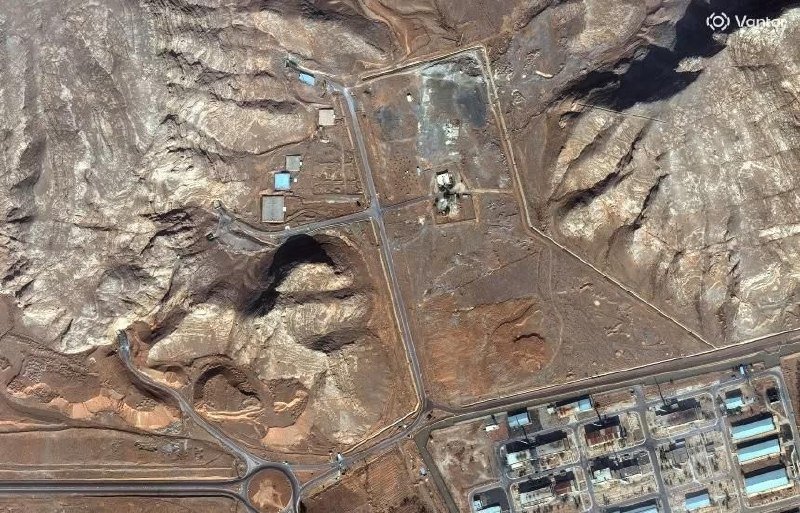 A satellite image shows a fenced compound with scattered buildings and tanks set in arid, rocky terrain, adjacent to an industrial facility with large structures.