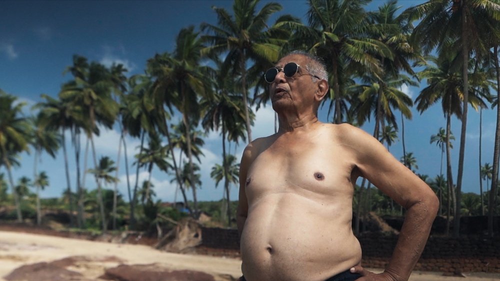 A shirtless older man wearing sunglasses stands on a tropical beach with tall palm trees lining the shoreline under a clear blue sky.