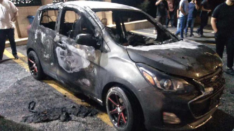 A burned-out hatchback car sits on a street at night, its windows shattered and interior charred, while several bystanders stand nearby observing the wreckage.
