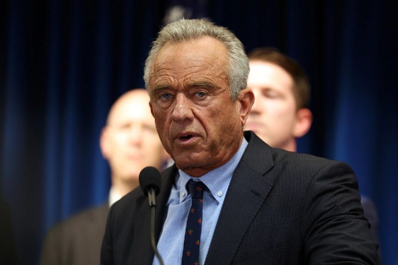 A gray-haired man in a dark suit and patterned tie speaks at a microphone, with two other men visible behind him against a blue curtain backdrop.