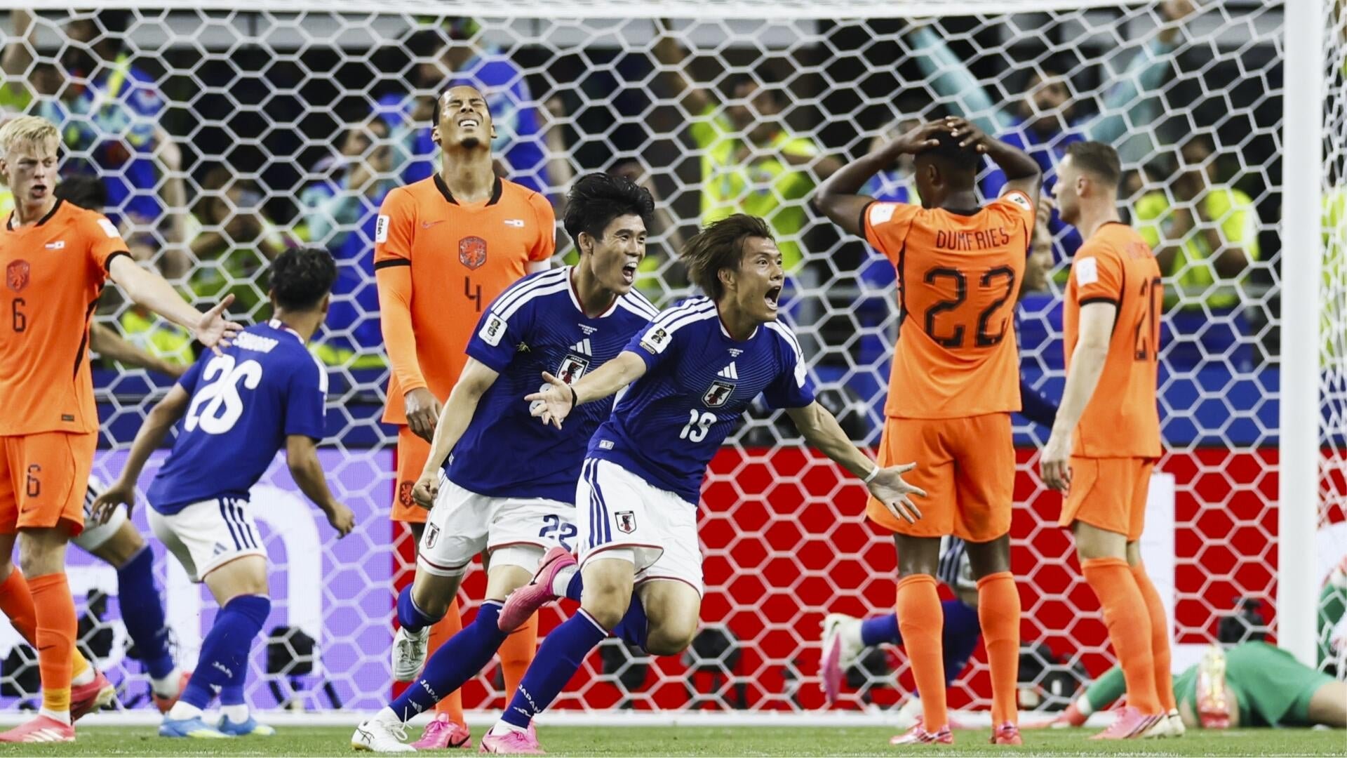 Soccer players in blue jerseys celebrate in front of the goal net while players in orange jerseys react with visible disappointment during a match.