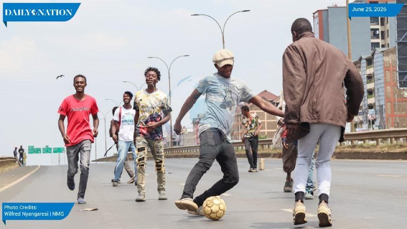 A group of young men play soccer on an urban roadway, with one dribbling a yellow ball while others run and challenge him.