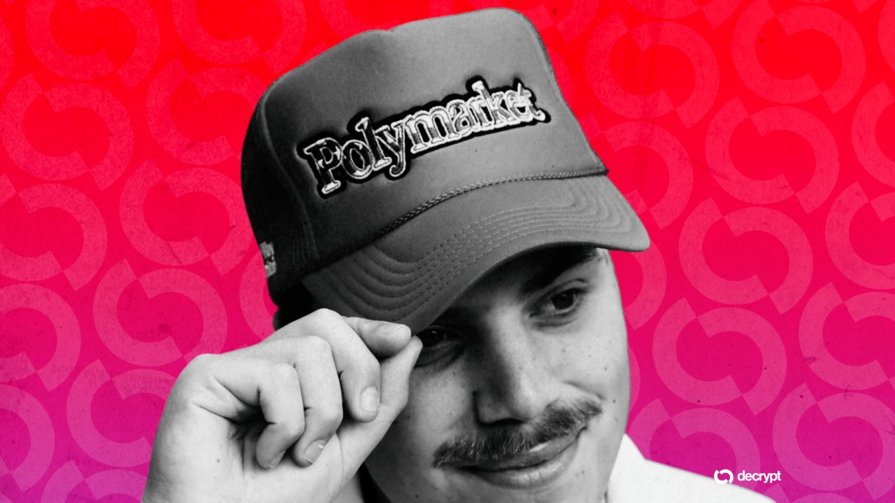 A man with a mustache tips a dark "Polymarket" baseball cap against a red patterned background with a "decrypt" logo in the corner.
