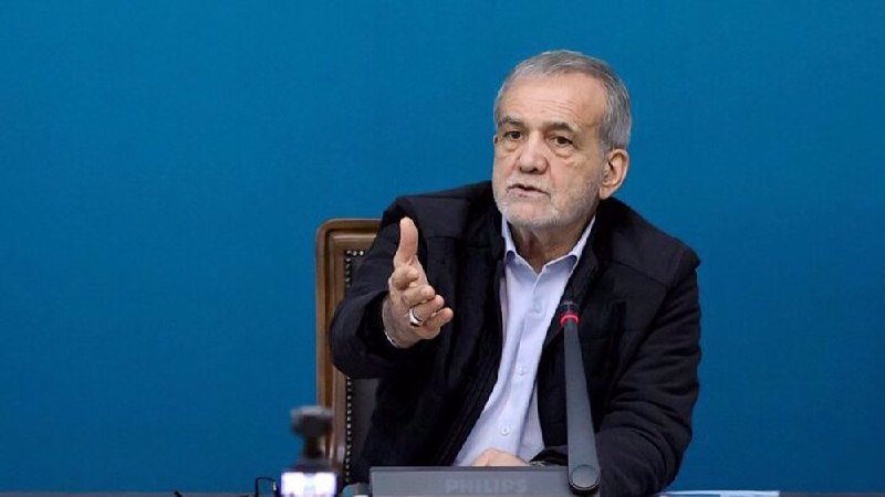 An older man with gray hair and a beard, wearing a black suit jacket and white shirt, gestures with his right hand while seated at a desk against a blue background.