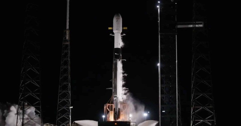 A white rocket lifts off from a launch pad at night, surrounded by tall support towers and billowing exhaust clouds.
