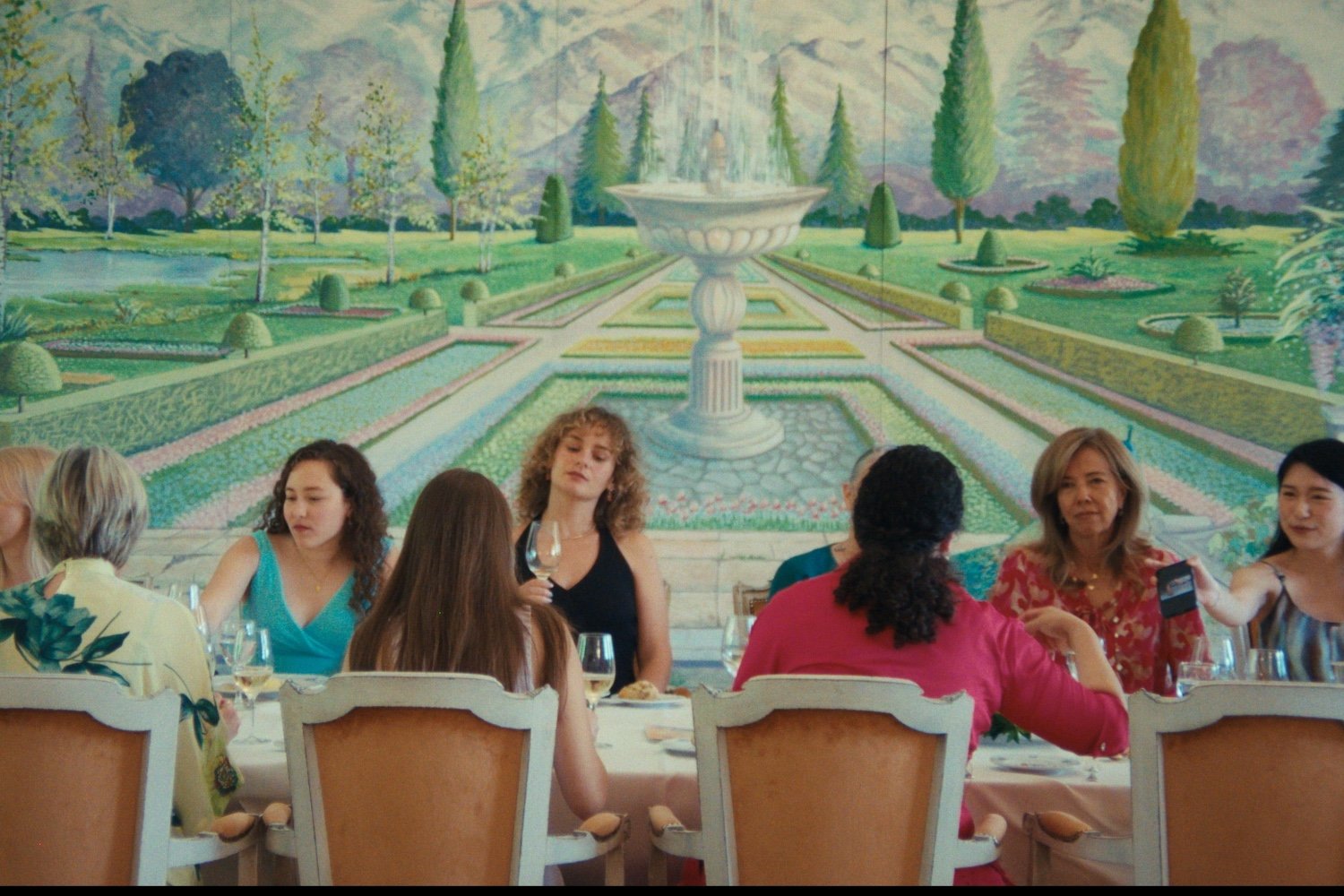Women sit at a dining table with wine glasses in front of an ornate mural depicting a formal garden with a fountain and mountains.