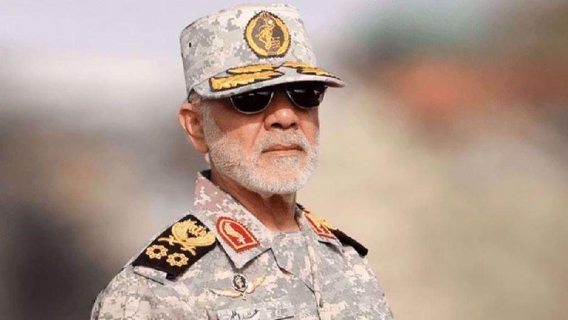 An older man with a white beard wearing a digital camouflage military uniform, cap with insignia, and sunglasses stands outdoors with a blurred background.