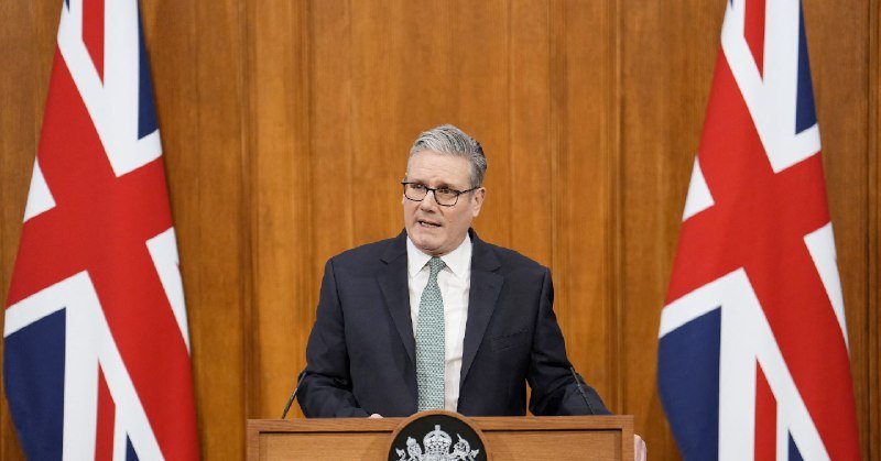 A bespectacled man in a dark suit and green tie stands at a wooden podium flanked by two Union Jack flags, appearing to speak.