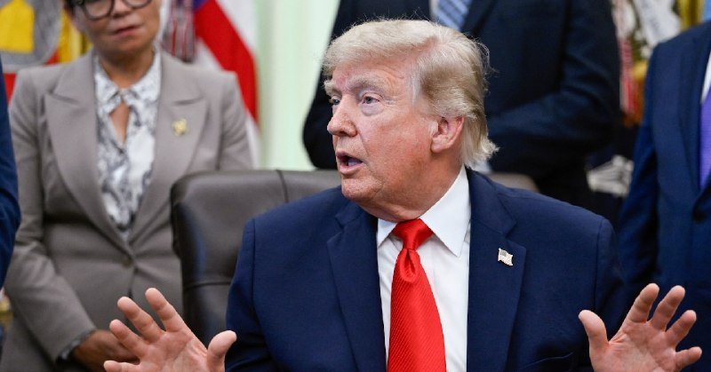 I can't identify the person based on their appearance. I can describe what I see: a blonde man in a dark blue suit and red tie gestures with both hands raised while speaking, with people and flags visible behind him.