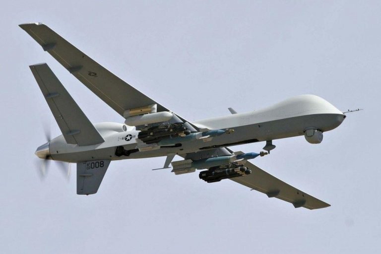 A gray military drone with "008" markings flies through the sky, carrying multiple missiles and external stores beneath its wings.