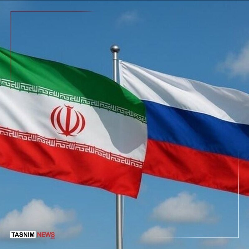 The Iranian flag and Russian flag wave side by side on a flagpole against a blue, partly cloudy sky.
