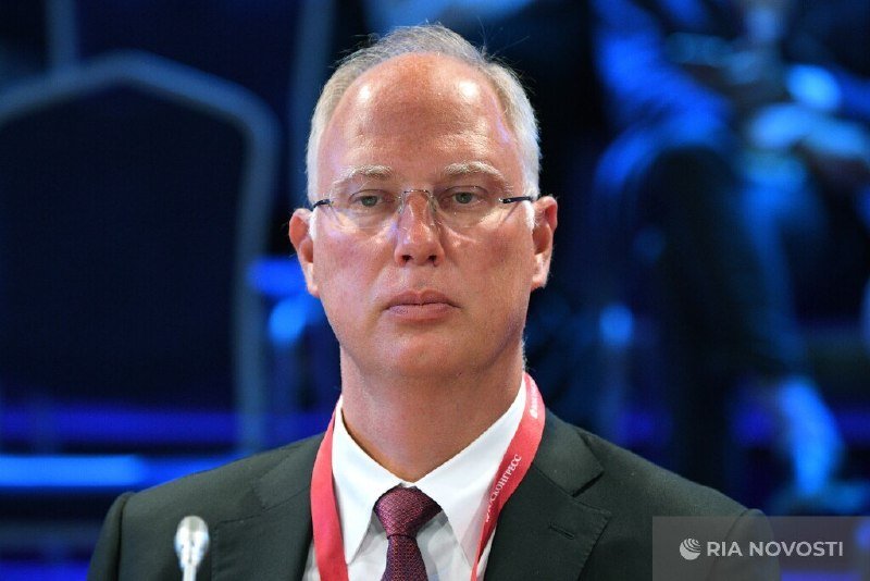 A bespectacled, balding man in a dark suit and red tie wears a lanyard labeled "NOVOTEC," standing before a blue-lit stage with the "RIA NOVOSTI" logo visible.