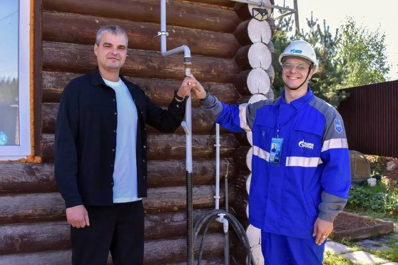 A man in a black jacket and a worker in a blue "Gazprom" uniform shake hands beside a log cabin with newly installed gas piping.