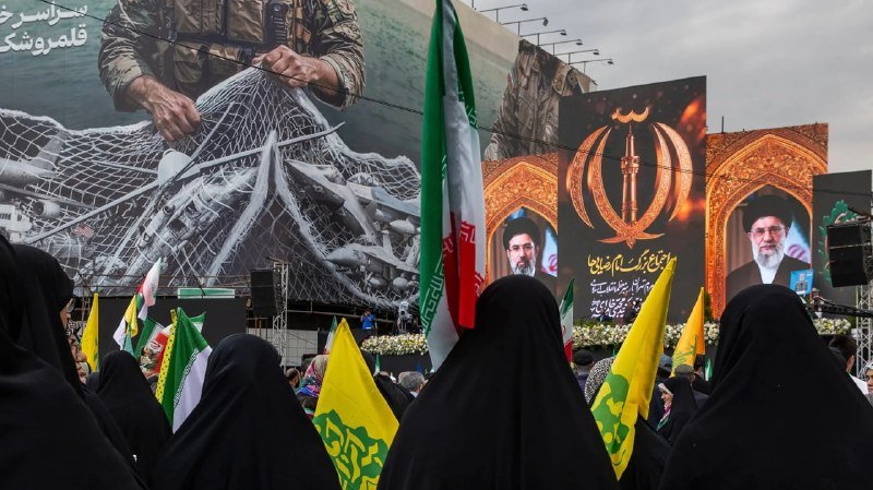 Women in black chadors holding Iranian and yellow Hezbollah flags gather before a large billboard depicting portraits of Iranian leaders and a soldier handling a net.