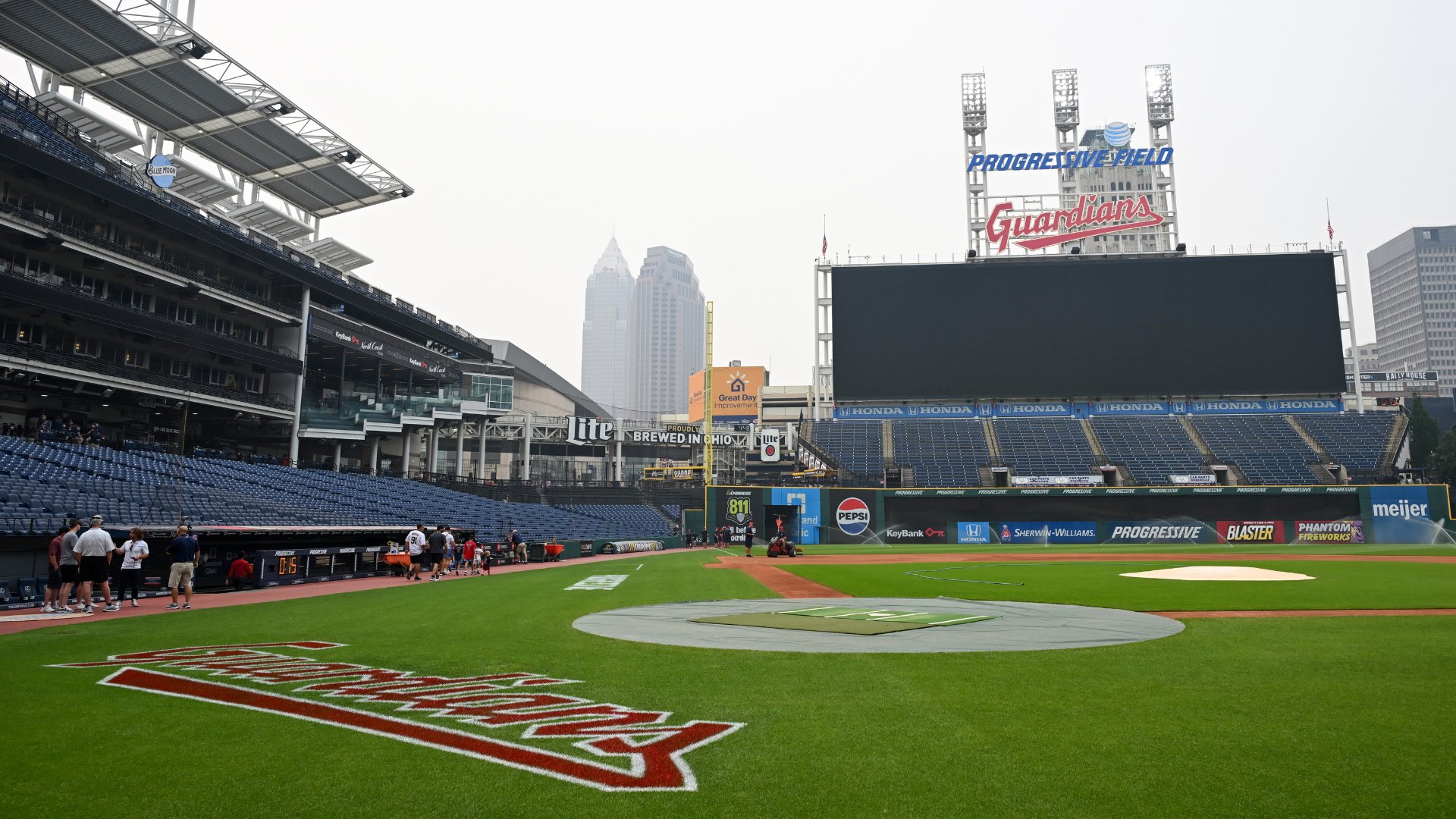 Empty baseball stadium with "Guardians" and "Progressive Field" signage, tarp covering home plate, overcast sky, and city skyline visible beyond the outfield.