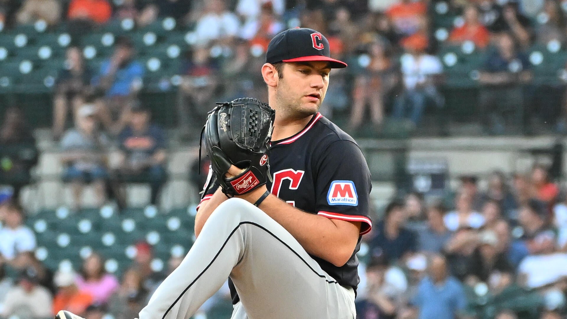 A baseball pitcher in a dark "C" cap and jersey winds up mid-pitch with his glove raised, one leg lifted, against a blurred crowd.