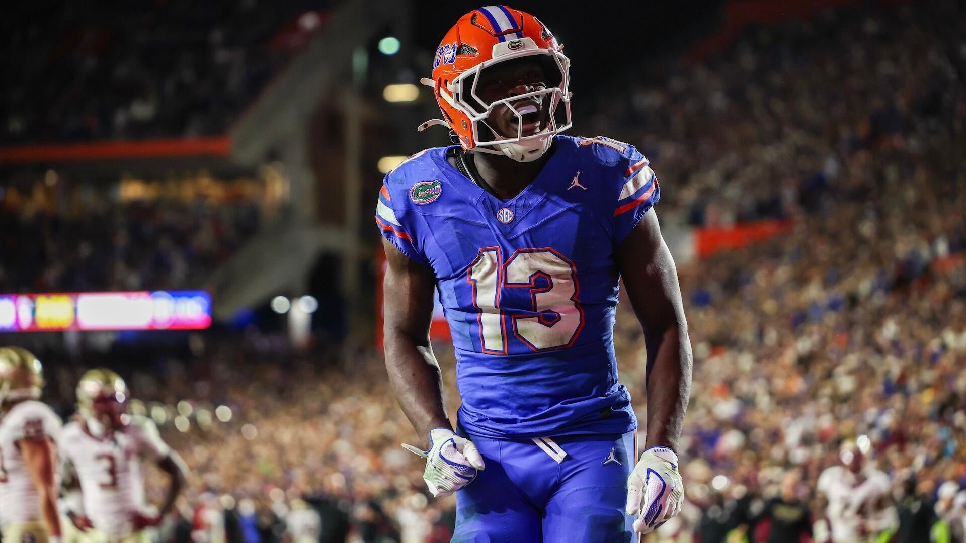 A Florida football player wearing jersey number 13 and an orange-and-blue helmet yells in celebration during a night game in a packed stadium.