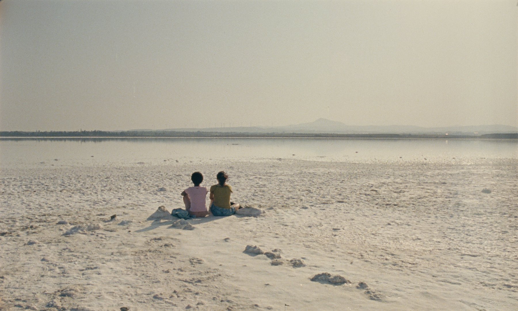 Two people sit side by side on a vast, white salt flat, gazing out at a calm body of water beneath a hazy sky with distant mountains.