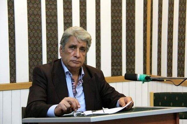 A gray-haired man in a dark blazer sits at a desk holding papers, speaking into a microphone against a striped wall.
