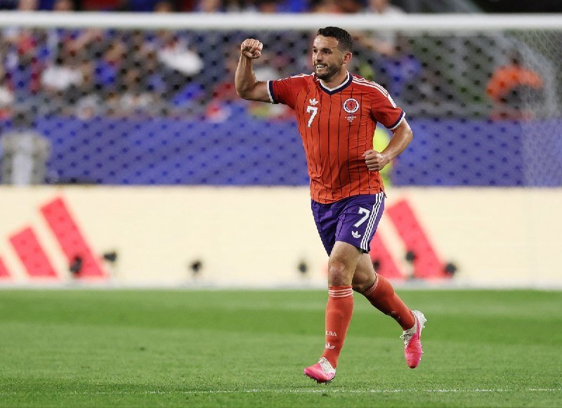 A soccer player in a red jersey and purple shorts, both bearing the number 7, celebrates with a raised fist on a stadium pitch in front of a blurred crowd.