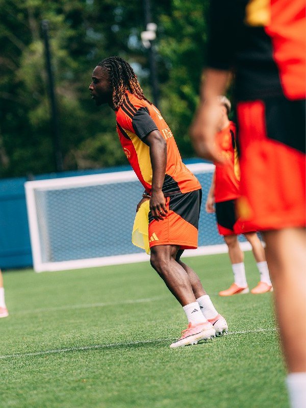 A soccer player with dreadlocks wearing an orange and black uniform holds a yellow pinnie on a green field during a training session.