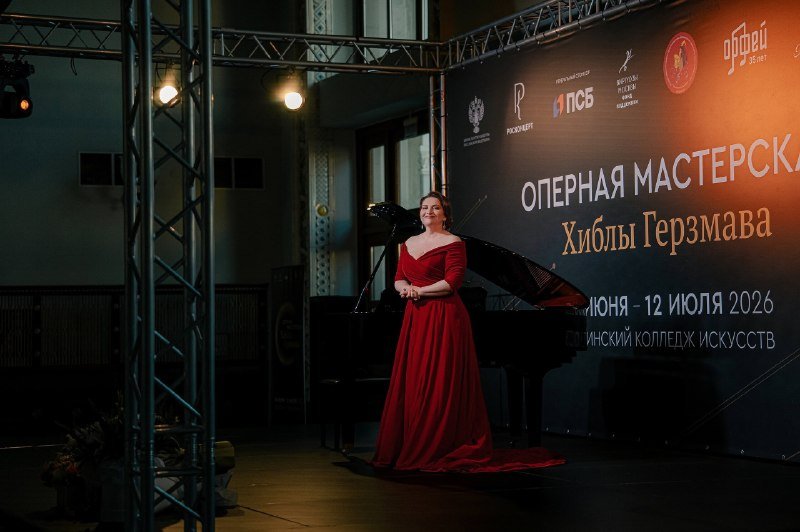A woman in a long red gown stands beside a grand piano on a lit stage, in front of a backdrop displaying Cyrillic text reading "ОПЕРНАЯ МАСТЕРСКАЯ Хиблы Герзмава."